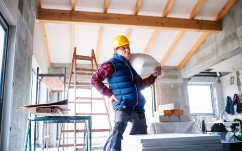 Image of a man at a work site rubbing his back as he carries a heavy sack 